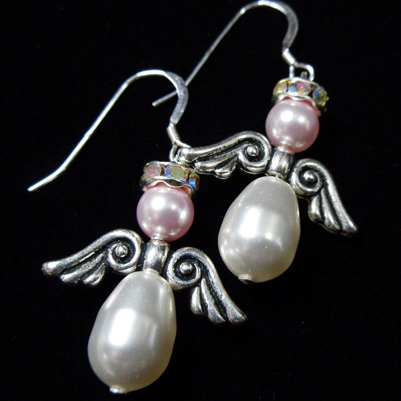 May include: A pair of silver-toned angel earrings with pink and white pearlescent beads. Each earring features a small pink bead at the top, a larger white bead, and silver wings. The earrings have a hook closure.