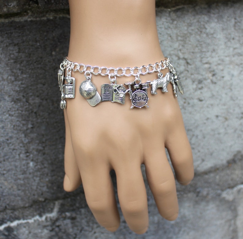 May include: A silver charm bracelet featuring a selection of charms, including a baseball cap, a book, a drum set, and a small elephant. The bracelet is worn on a person's wrist.