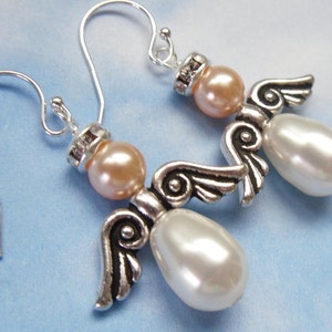 May include: A pair of angel-shaped earrings. Each earring features a peach-colored bead head, a silver-toned wing design, and a large, pearly white teardrop-shaped body. The earrings have silver-colored ear wires and are set against a light blue background.