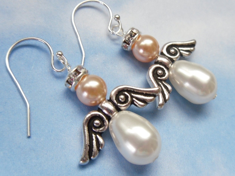 May include: A pair of angel-shaped earrings. Each earring features a peach-colored bead head, a silver-toned wing design, and a large, pearly white teardrop-shaped body. The earrings have silver-colored ear wires and are set against a light blue background.