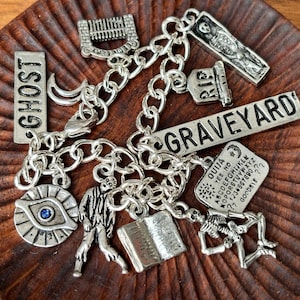 May include: A silver charm bracelet with various Halloween-themed charms, including a ghost, a graveyard, a Ouija board, a skeleton, and an eye. The charms are attached to a silver chain.