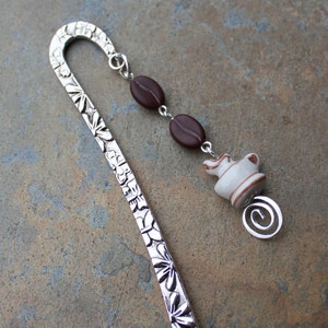 Op de afbeelding: Zilveren bladwijzer met een koffieboonontwerp en een bedel van een koffiepot met bruine koffiebonen en een zilveren werveling.