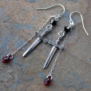 Pode incluir: Brincos de espada prateados com detalhes em cristal preto e vermelho. As espadas são decoradas com desenhos intrincados e penduradas em correntes delicadas.