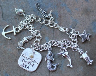May include: Silver charm bracelet with a variety of sea-themed charms, including a mermaid, a dolphin, a starfish, an anchor, a seashell, and a seahorse. The bracelet also features a charm with the text "Dreaming of the Sea".