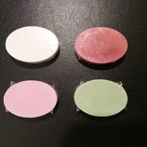 May include: Four oval-shaped bars of soap wrapped in clear plastic. The bars are colored white, reddish-pink, light pink, and pale green. Each bar has a smooth, solid surface and is arranged in a two-by-two grid pattern on a dark surface.