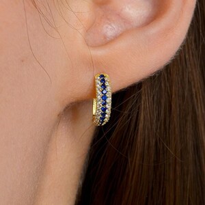 May include: A small gold hoop earring featuring a central row of deep blue stones flanked by two rows of clear, sparkling stones. The hoop is circular in shape and designed to sit snugly against the earlobe, showcasing a polished gold finish.
