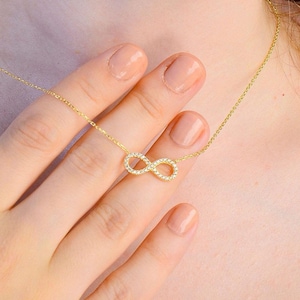 May include: A gold-toned necklace featuring a central infinity symbol pendant encrusted with small, clear, sparkling stones. The delicate chain rests around the neck, showcasing the horizontal figure-eight design that catches the light.