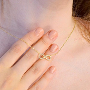 May include: A gold necklace with a small infinity symbol pendant encrusted with tiny clear stones. The necklace is worn by a person with their hand resting on their chest.