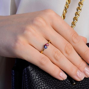 May include: A gold ring featuring two heart-shaped gemstones, one red and one blue. The ring is set on a thin gold band. The ring is displayed on a hand, with a black leather purse in the background.