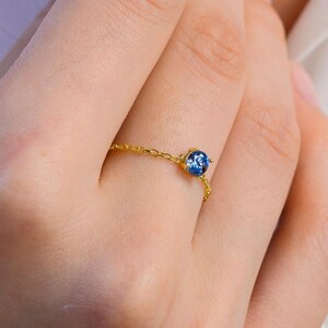 May include: A delicate gold chain ring featuring a single, small, round, faceted blue gemstone set in a gold prong setting. The ring band is constructed from fine gold chain links that wrap around the finger, showcasing a minimalist design.