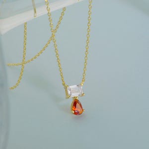 May include: A gold chain necklace featuring a horizontal emerald-cut clear stone pendant with a smaller, dangling pear-shaped orange stone. The delicate metal links support the two-stone setting, creating a refined and balanced jewelry piece.
