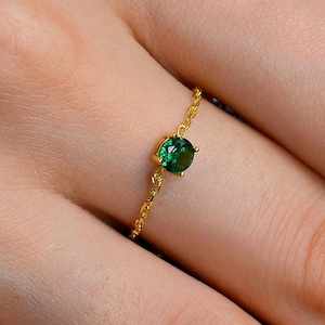 May include: A delicate gold chain ring featuring a single round-cut emerald green gemstone set in a four-prong setting. The band is constructed from fine gold links, creating a flexible and lightweight appearance that encircles the finger.