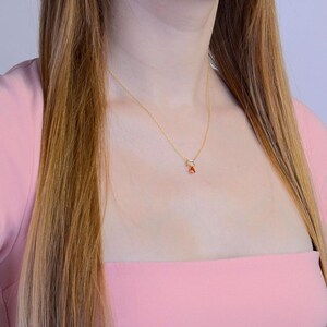 May include: A delicate gold chain necklace featuring a small pendant with a clear, rectangular-cut stone positioned above a vibrant, orange, pear-shaped gemstone. The necklace rests against the neck, showcasing a minimalist and refined design.