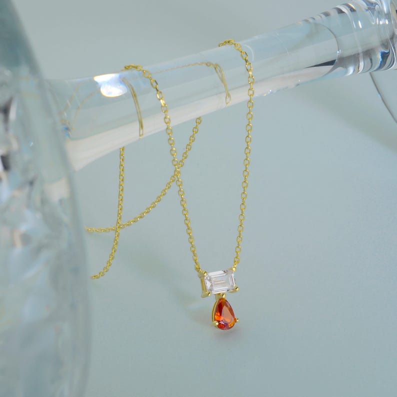 May include: A gold chain necklace featuring a horizontal emerald-cut clear stone pendant with a smaller, dangling pear-shaped orange stone. The delicate metal links support the two-stone setting, creating a refined and balanced jewelry piece.