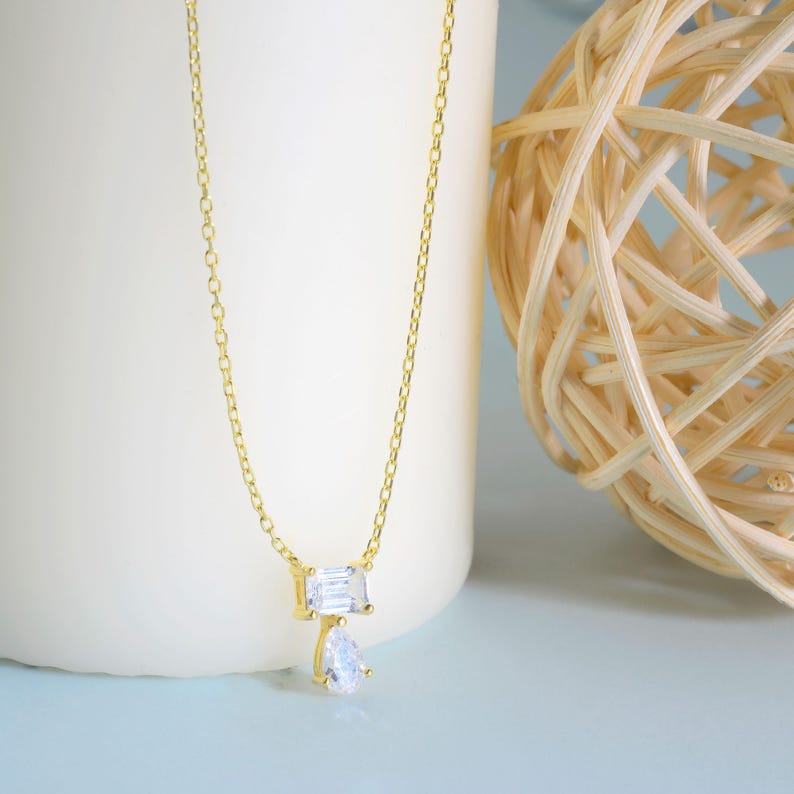 May include: A gold-toned chain necklace featuring a pendant with a rectangular baguette-cut clear stone positioned above a pear-shaped clear stone. The metal chain is thin and delicate, connecting to the two stones set in a gold-coloured metal frame.