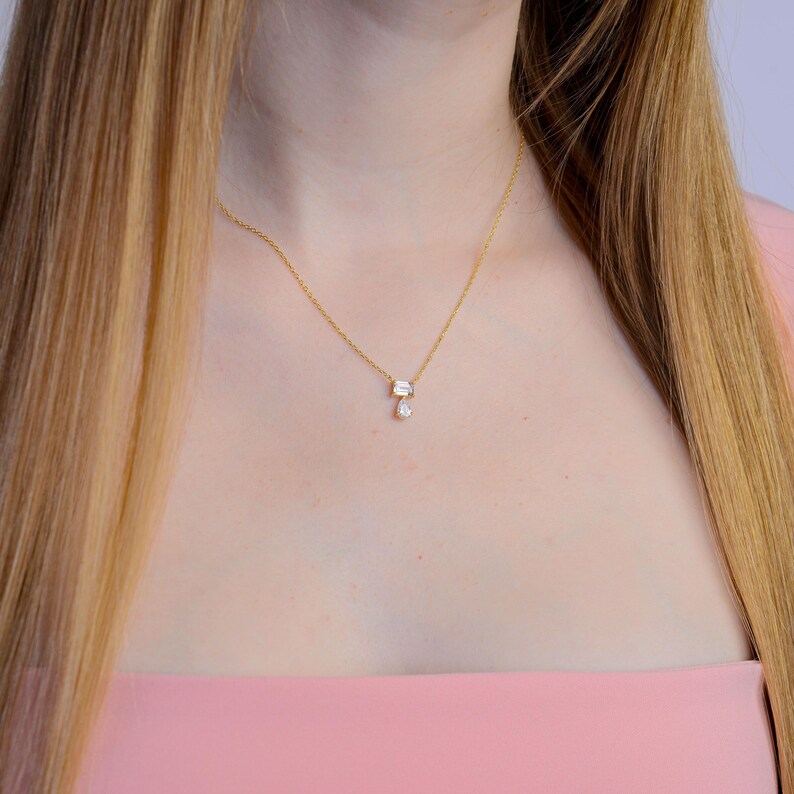 May include: A gold-toned chain necklace featuring a pendant with a rectangular baguette-cut clear stone positioned horizontally above a smaller, dangling pear-shaped clear stone. The necklace rests against the neck, showcasing a delicate metal link design.