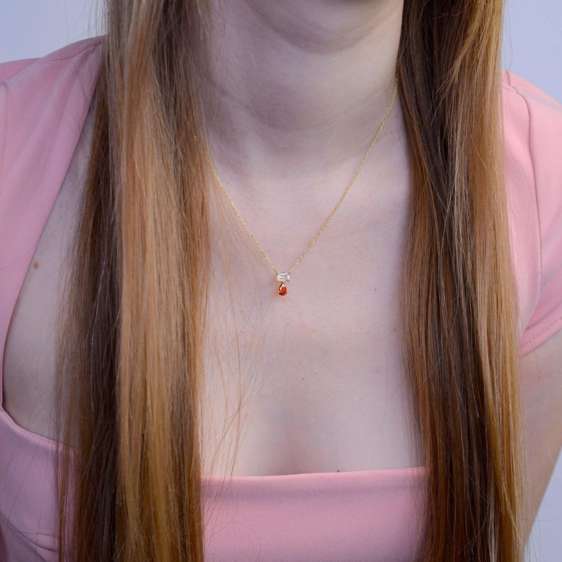 May include: A delicate gold chain necklace featuring a small pendant with a clear rectangular stone positioned above a vibrant orange teardrop-shaped gemstone. The necklace rests against the neck, showcasing a minimalist design with two distinct stones.