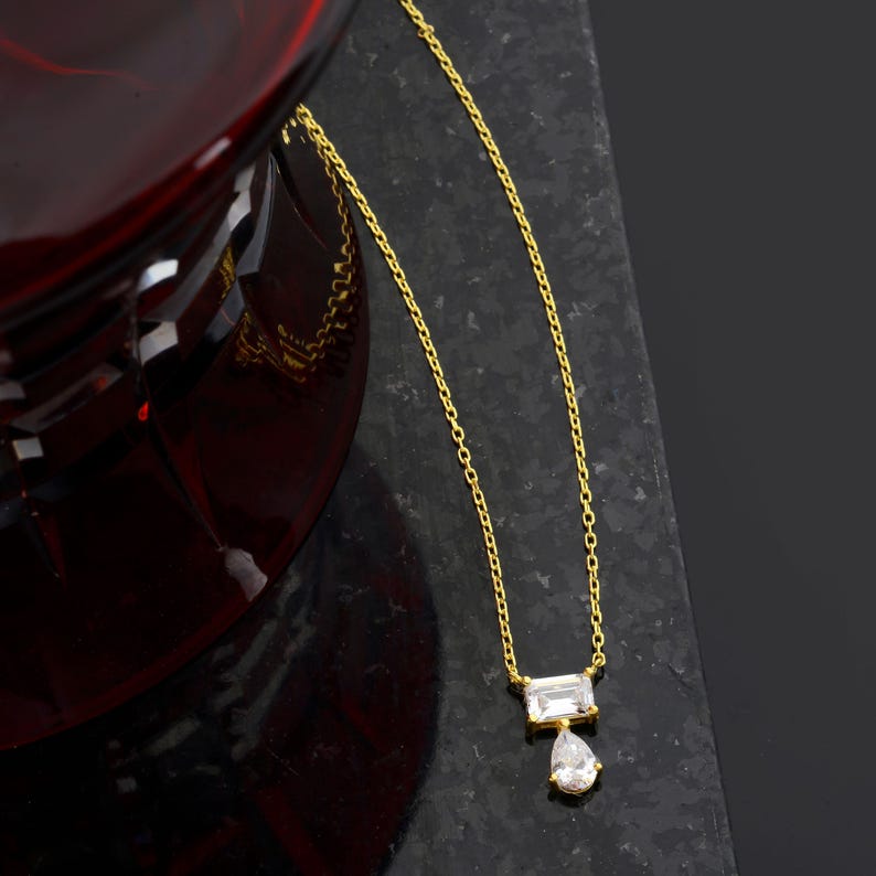 May include: A gold-toned necklace featuring a delicate chain with a pendant composed of a rectangular baguette-cut clear stone positioned horizontally above a smaller, dangling pear-shaped clear stone, creating a layered geometric design.