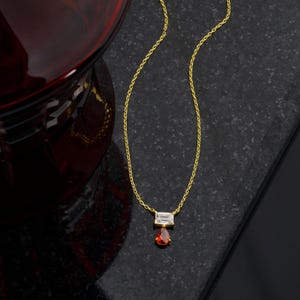 May include: A gold-toned chain necklace featuring a horizontal baguette-cut clear stone pendant with a dangling pear-shaped red stone drop. The delicate metal chain connects to the rectangular stone setting, which supports the vibrant red teardrop gem.