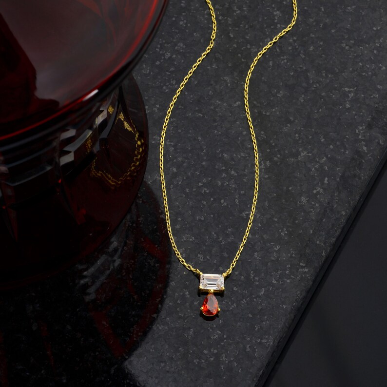 May include: A gold-toned chain necklace featuring a horizontal baguette-cut clear stone pendant with a dangling pear-shaped red stone drop. The delicate metal chain connects to the rectangular stone setting, which supports the vibrant red teardrop gem.