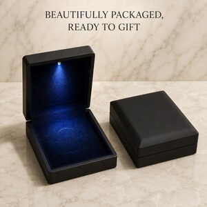 May include: Two rectangular black jewellery boxes with matte finishes. One box is open, revealing a dark blue velvet interior illuminated by a small, bright LED light at the top. The text "BEAUTIFULLY PACKAGED, READY TO GIFT" is visible above the boxes.