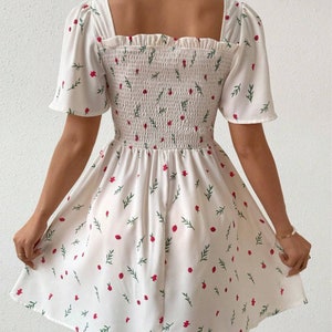 May include: White summer dress with a floral print of red flowers and green leaves. The dress has a square neckline, short puff sleeves, and a smocked bodice.