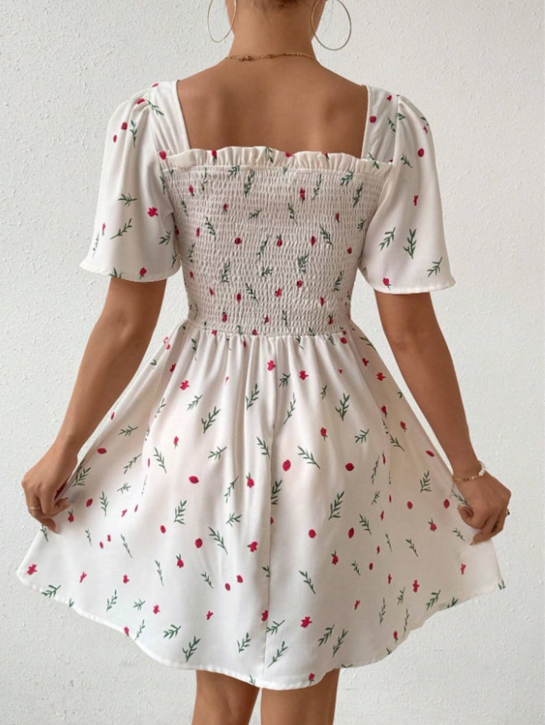 May include: White summer dress with a floral print of red flowers and green leaves. The dress has a square neckline, short puff sleeves, and a smocked bodice.