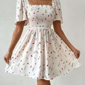 May include: A white mini dress with a square neckline and short puff sleeves. The dress has a floral print with red flowers and green leaves.