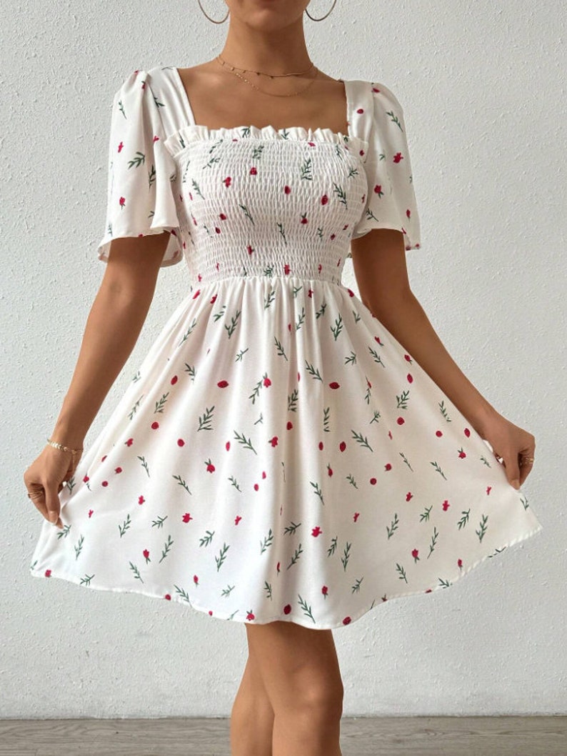 May include: A white mini dress with a square neckline and short puff sleeves. The dress has a floral print with red flowers and green leaves.