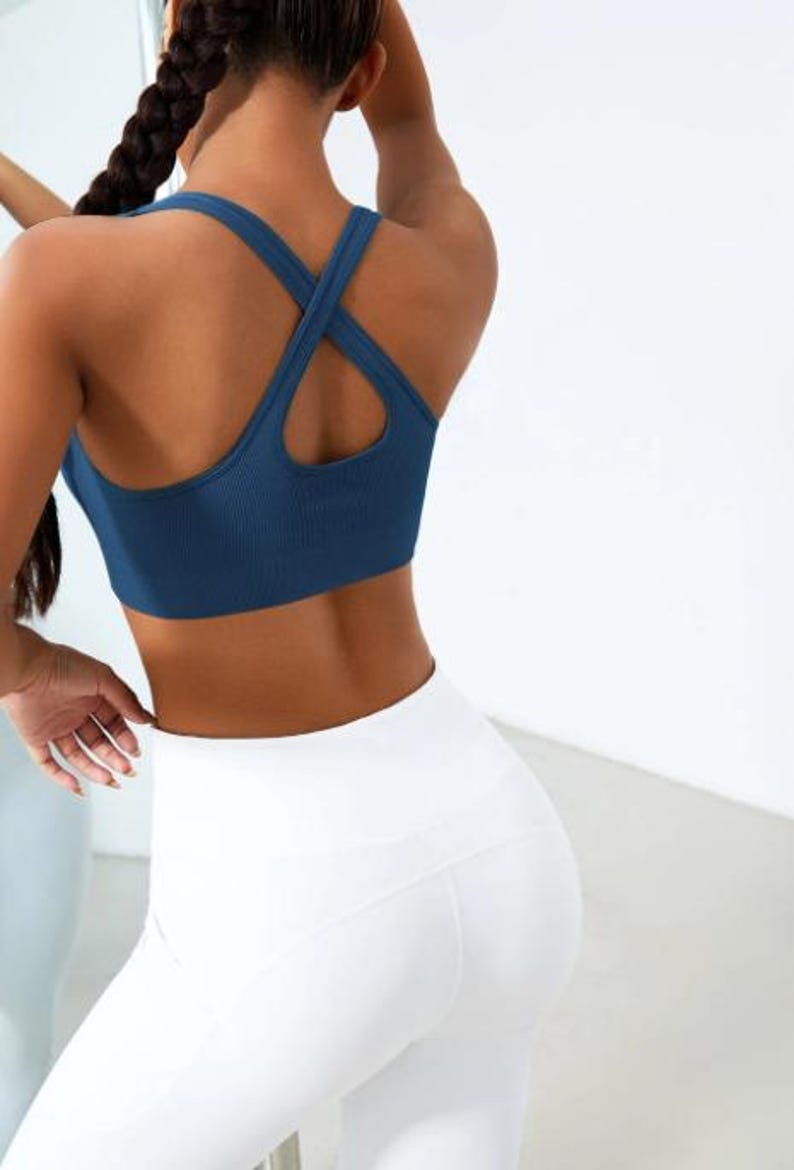 May include: A navy blue sports bra with a crisscross back design is paired with white leggings. The bra has a ribbed texture. The leggings are high-waisted and form-fitting. The image is taken from the back, showcasing the athletic wear.