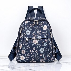 May include: A navy blue backpack featuring a floral pattern with light pink and pale blue blossoms, green leaves, and thin stems. The bag includes silver-toned metal zippers, a top handle, and adjustable shoulder straps in a solid navy blue.