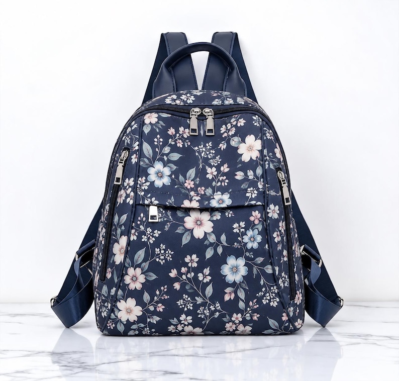 May include: A navy blue backpack featuring a floral pattern with light pink and pale blue blossoms, green leaves, and thin stems. The bag includes silver-toned metal zippers, a top handle, and adjustable shoulder straps in a solid navy blue.