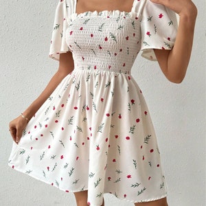 May include: White summer dress with a floral print of red flowers and green leaves. The dress has a square neckline, short puff sleeves, and a fitted bodice. The skirt is flowy and has a smocked waist.