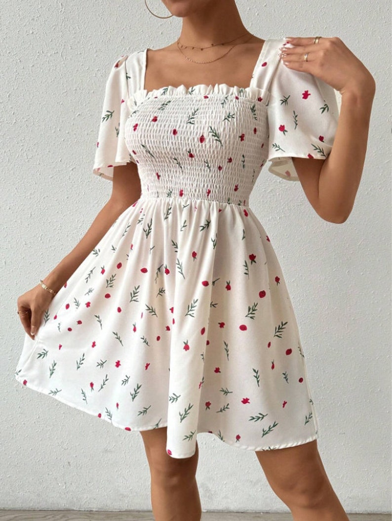 May include: White summer dress with a floral print of red flowers and green leaves. The dress has a square neckline, short puff sleeves, and a fitted bodice. The skirt is flowy and has a smocked waist.
