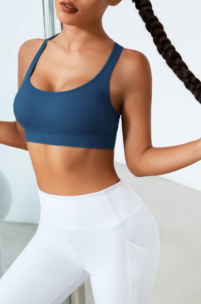 May include: A woman wearing a teal blue ribbed sports bra and white leggings. The sports bra has a scoop neck and wide straps. The leggings have a high waist and pockets. The woman has long dark hair.