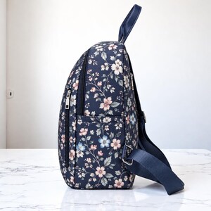 May include: A navy blue backpack featuring a floral pattern with light pink, white, and pale blue flowers and green leaves. The bag includes a top handle, adjustable navy blue shoulder straps, and a side zipper pocket with silver hardware.