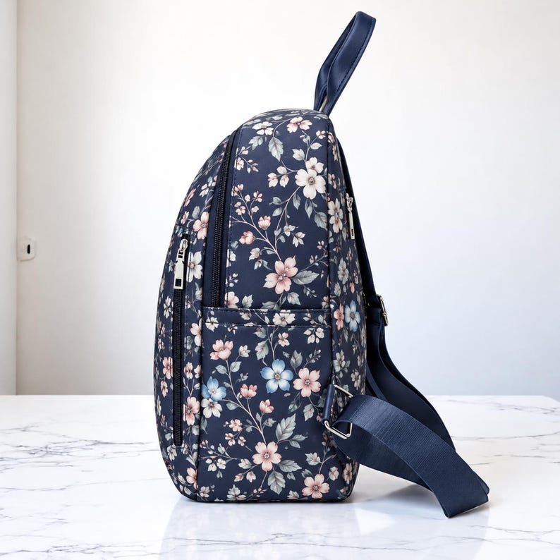May include: A navy blue backpack featuring a floral pattern with light pink, white, and pale blue flowers and green leaves. The bag includes a top handle, adjustable navy blue shoulder straps, and a side zipper pocket with silver hardware.