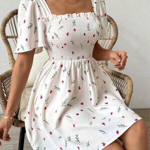May include: White summer dress with a floral print featuring small red flowers and green leaves. The dress has a square neckline, short puff sleeves, and a fitted bodice with a smocked elastic waistband. The skirt is flowy and falls to just above the knee.