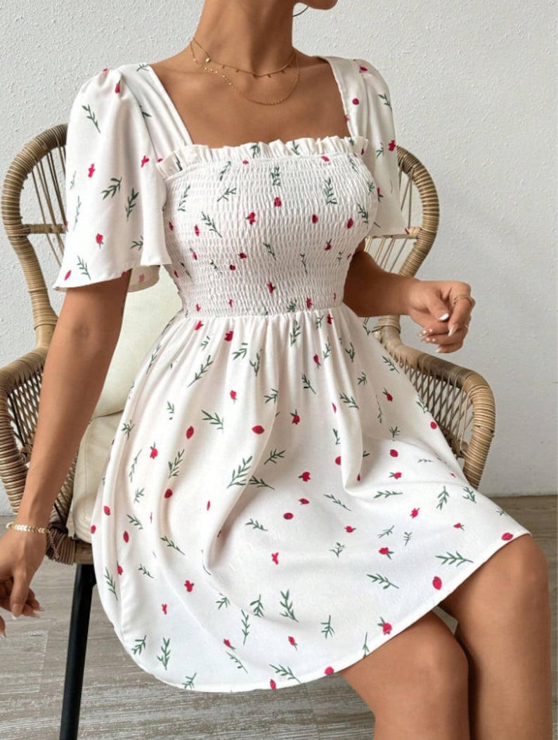 May include: White summer dress with a floral print featuring small red flowers and green leaves. The dress has a square neckline, short puff sleeves, and a fitted bodice with a smocked elastic waistband. The skirt is flowy and falls to just above the knee.