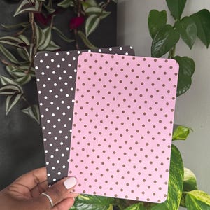 May include: Two rectangular cards with rounded corners. One card is dark grey with a white polka dot pattern. The second card is light pink with a brown polka dot pattern. Both cards are held together in a vertical orientation.