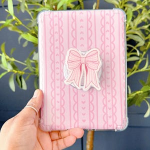 May include: A pink tablet case featuring a bow-shaped pop socket. The case has a vertical striped pattern with heart and scalloped details. The pop socket is a light pink bow with a white background. The case is held in a clear plastic protective cover.