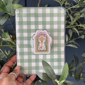 Op de afbeelding: Een transparante beschermhoes voor een tablet, met een groen en wit ruitpatroon. Een decoratieve sticker met een sleutelontwerp en een roze strik is aan de hoes bevestigd. Het geheel is omgeven door groen gebladerte.