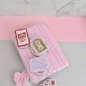 May include: A pink phone case with a repeating pattern, adorned with several decorative pop sockets. The pop sockets feature various designs, including a bow, a key, and text that reads "Buy Me Books" and "Meet me at the Bookshop".