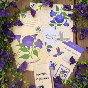 May include: A collection of stationery items featuring purple morning glory flowers and hummingbird illustrations, including cards with the text Morning Glory, Lavender Hour, September, and September in pictures, surrounded by artificial purple flowers.