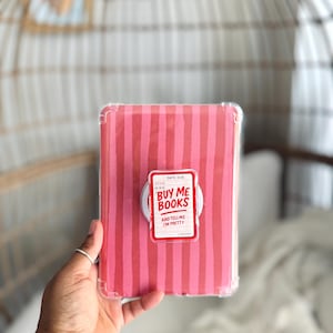 May include: A pink and red striped book cover with a white pop socket. The pop socket has a red and white sticker that says "BUY ME BOOKS AND TELL ME I'M PRETTY". The book cover is in a clear case.