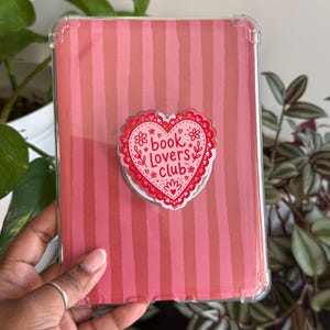 May include: A clear protective case for an e-reader with a pink striped design. A heart-shaped pop socket with the words "book lovers club" is attached to the case. The pop socket has a red and white floral design.