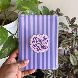 以下が含まれることがあります： 紫色のストライプ模様の透明な保護ケース。 「Books and Coffee」という言葉が入ったピンクと白のステッカーが貼られています。 ケースは緑の植物の前に置かれています。