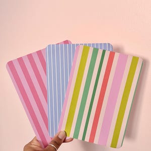 May include: Three rectangular, striped decorative items in shades of pink, blue, green, and yellow. The items are held against a light pink background. The stripes are vertical and vary in width and colour.