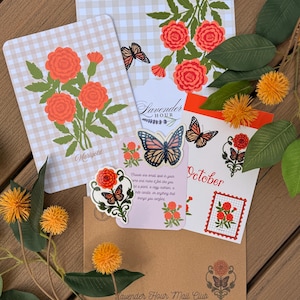 May include: A collection of stationery items including a card with orange marigolds on a beige gingham background, a sticker sheet labelled October, a small card with text, and a brown envelope stamped with Lavender Hour Mail Club.