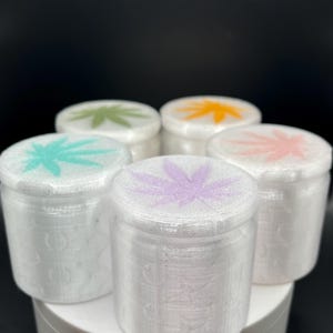 May include: A collection of six white, cylindrical containers with lids. Each lid features a cannabis leaf design in a different color: green, orange, pink, purple, and teal. The containers are arranged on a white platform against a black background.